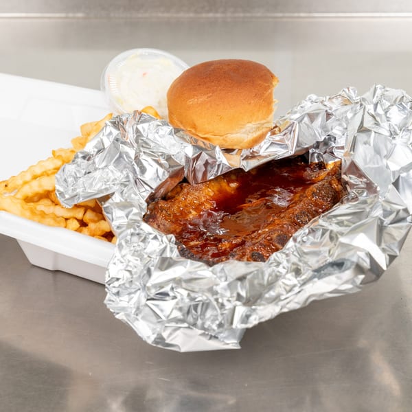 Ribs with BBQ sauce, crinkle-cut fries, a bun, and coleslaw in a white takeout container.