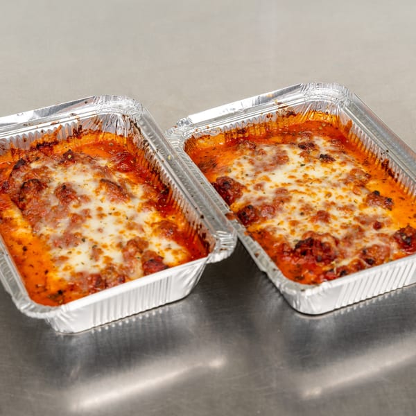 Two aluminum foil trays filled with baked ziti, topped with melted cheese and tomato sauce.