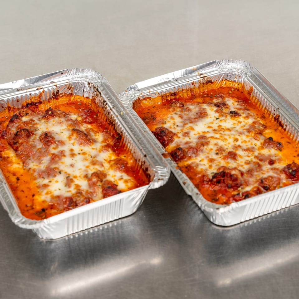 Two aluminum foil trays filled with baked ziti, topped with melted cheese and tomato sauce.