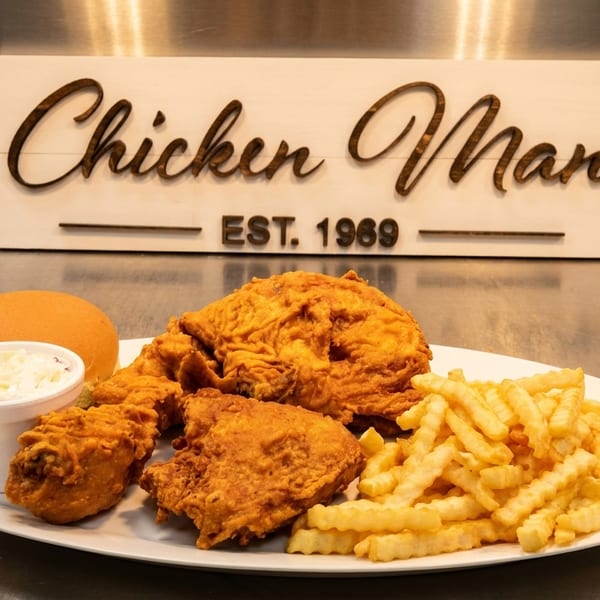 A plate of Chicken Man fried chicken, crinkle-cut fries, coleslaw, and a bun on a metal counter.