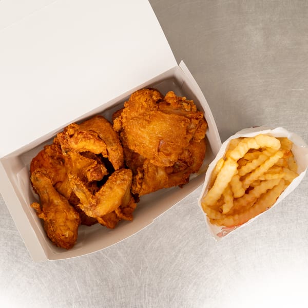 Crispy fried chicken pieces in a white takeout box next to a bag of crinkle-cut fries.