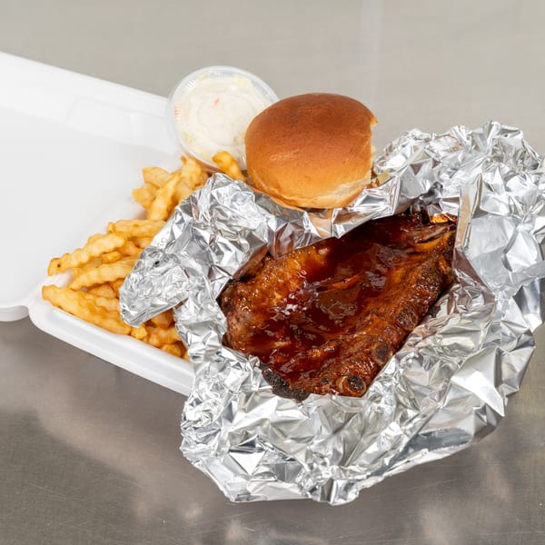 BBQ ribs in foil with fries, coleslaw, and a bun in a white takeout container.