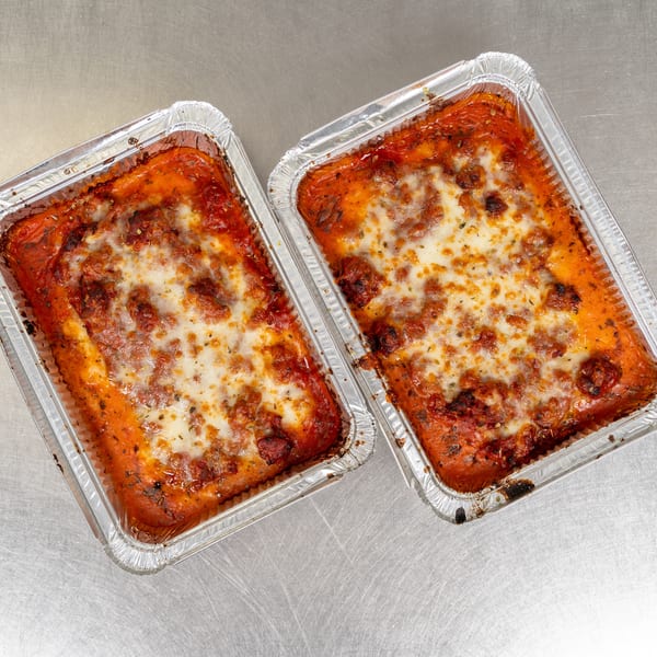 Two aluminum foil trays of baked lasagna with melted cheese and tomato sauce on a metal surface.