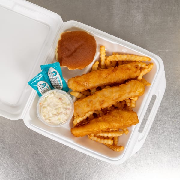 A takeout container holds golden-brown fried fish, crinkle-cut fries, a bun, coleslaw, and tartar sauce packets.